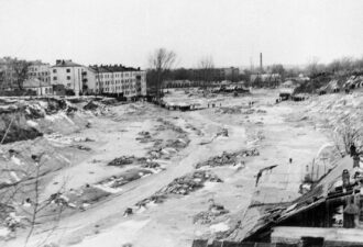 Чорно-біла фотографія зображує наслідки масштабної повені в міському районі. На передньому плані видно зруйнований дерев’яний будинок із пошкодженим дахом. Посеред кадру – русло висохлої бурхливої води, заповнене уламками та багнюкою. Ліворуч розташовані багатоповерхові житлові будинки, біля яких зібралися люди. На задньому плані видно промислові будівлі, труби заводів і довгі черги людей, що вишикувалися на пагорбах, спостерігаючи за ситуацією. Загальний вигляд створює атмосферу руйнування та втрат після стихійного лиха.