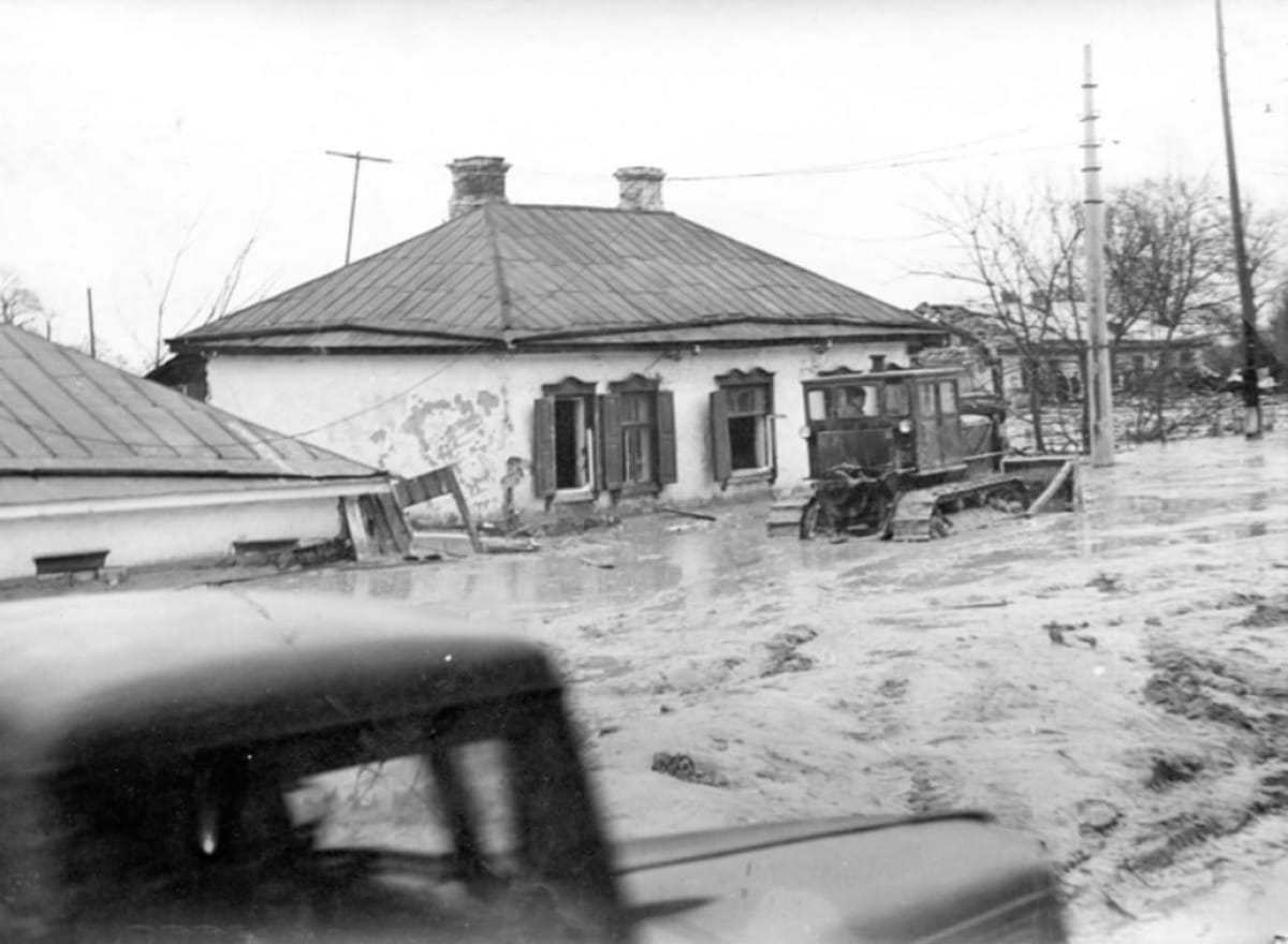 Чорно-біла фотографія показує наслідки повені в населеному пункті. Одноповерховий будинок із металевим дахом частково затоплений, а стіни виглядають пошкодженими. Біля будівлі застряг гусеничний трактор, наполовину занурений у воду та багнюку. На передньому плані видно розмитий силует автомобіля, що рухається. Навколо спустошений пейзаж, поламані конструкції та підтоплені дерева, що свідчать про масштаби стихійного лиха.
