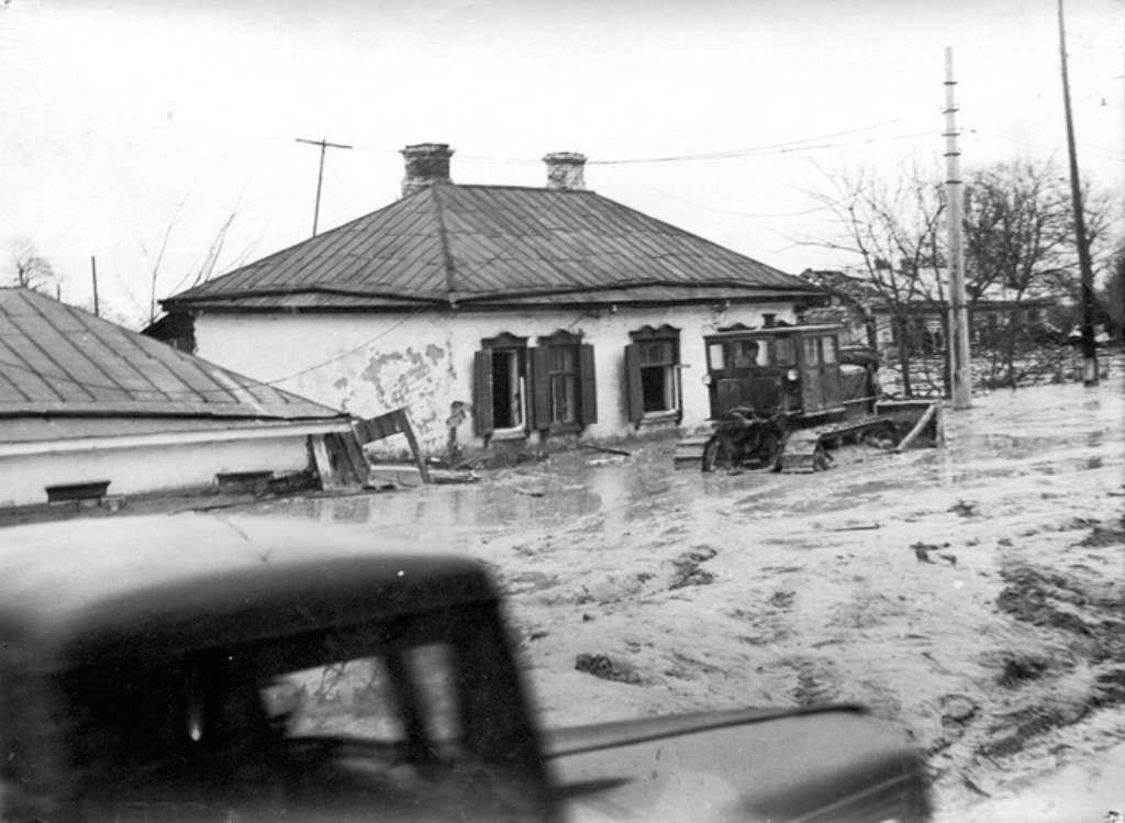 Чорно-біла фотографія показує наслідки повені в населеному пункті. Одноповерховий будинок із металевим дахом частково затоплений, а стіни виглядають пошкодженими. Біля будівлі застряг гусеничний трактор, наполовину занурений у воду та багнюку. На передньому плані видно розмитий силует автомобіля, що рухається. Навколо спустошений пейзаж, поламані конструкції та підтоплені дерева, що свідчать про масштаби стихійного лиха.