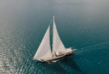Білий вітрильник із двома щоглами пливе бірюзовими водами моря під британським прапором, знято з висоти.