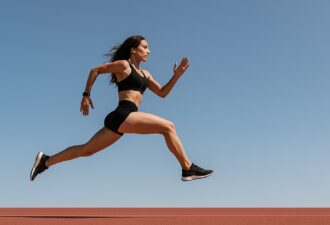 Спортивна дівчина в стрибку на біговій доріжці, на фоні блакитного неба, в яскравому денному світлі.